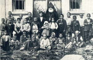 A group of rescued orphans standing together with an Armenian priest. The background is a building with one door and two windows.