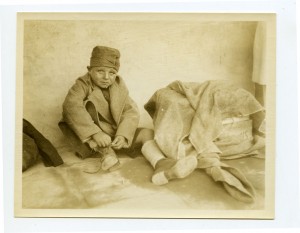 A small boy tying his shoe, wearing a coat and a hat and his eyes are looking up.