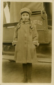 Girl in winter clothes standing in front of truck with Near East Relief insignia on the door.