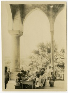 Children take their meal at a long table on the colonnaded porch of the Birds' Nest Orphanage. Nellie Miller's original caption reads 