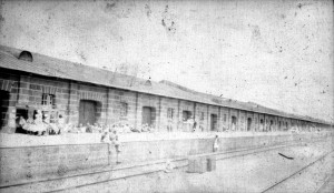 A Garage along train tracks in Kazachi from Jaquith collection. Many people standing on the side of the train tracks with their pieces of luggage and belongings.