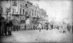 Photograph of a city street with soldiers visible in the middle ground. The soldiers are wearing uniforms and carrying rifles. The image is damaged, and the date and location are unknown. It is possible that the soldiers are members of a Greek army battalion either entering or leaving the city of Smyrna.