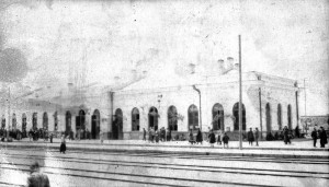 View of train station, most likely in Kars, Turkey. Numerous train tracks are visible in the foreground. The building is a long one-story structure with prominent arched windows in the Roman style. The date is unknown.