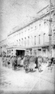 Young men in uniform on a city street in front of a large building. The young men may be dressed as Boy Scouts, or it may be an orphanage uniform. They wear matching shirts, shorts, knee socks, and hats. The boys in the second row appear to be carrying a small trunk. There is a small crowd gathered on the sidewalk, indicating that the boys may be marching in a parade.