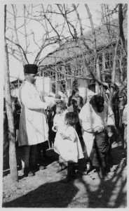 A man in a distinctive round black hat and white coat, and another adult in a similar outfit clip the hair of young children. The adults hold shears. There are tufts of hair visible on the ground. New arrivals at orphanages usually had their heads shaved to prevent the spread of lice (which carried typhus) and the contagious scalp disease favus. Probably Tiflis, c. 1920.