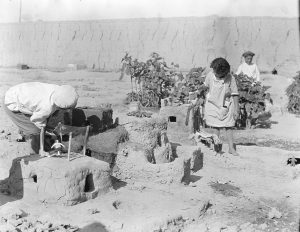 A boy bends to inspect a small house built from mud bricks in the garden at Alexandropol. The children enjoyed building and decorating these playhouses with readily available natural materials. The house at left has the additional luxury of a small doll.