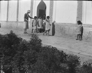 Girls in aprons walking out of an orphanage building while a relief worker guides them. Although the location is unknown, a girl in a similar apron and dress appears in a photograph from Alexandropol.