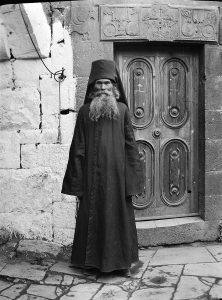 An Eastern Orthodox priest in clerical garb standing in front of a church door. The headpiece indicates that the priest is Russian rather than Armenian.