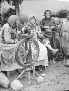 A group of women gathered around a spinning wheel. Two of the women are knitting. The ability to spin and knit helped women to be more self-sufficient.