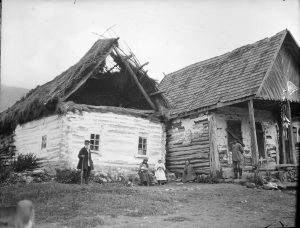 Three women sit in front of two dilapidated buildings. One woman holds a child. The woman in the center might be dressed as a nurse. There are two men standing on either side.