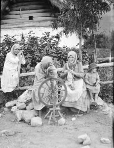 Two women sit at a spinning wheel. One woman knits. Two children look on. 
